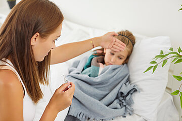 Eine Mutter sitzt bei ihrer kleinen Tochter am Krankenbett und f&uuml;hlt die Stirn.