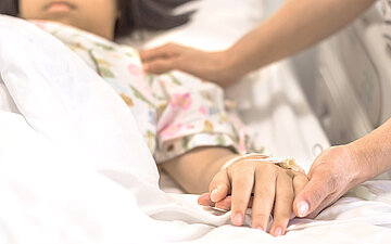 Foto: todkrankes M&auml;dchen liegt im Krankenhausbett, Erwachsene h&auml;lt ihre Hand.