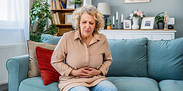 Eine Frau sitzt im Wohnzimmer auf einer hellblauen Couch und h&auml;lt sich mit schmerzverzerrtem Gesicht den Bauch.