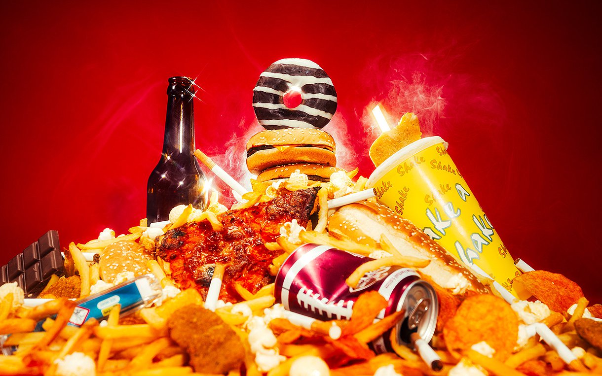 Ungesunde Lebensmittel, wie Pommes, Donuts, Alkohol, Zigaretten, Süßkram etc. liegen vergoldet und scheinend auf einem großen Haufen.