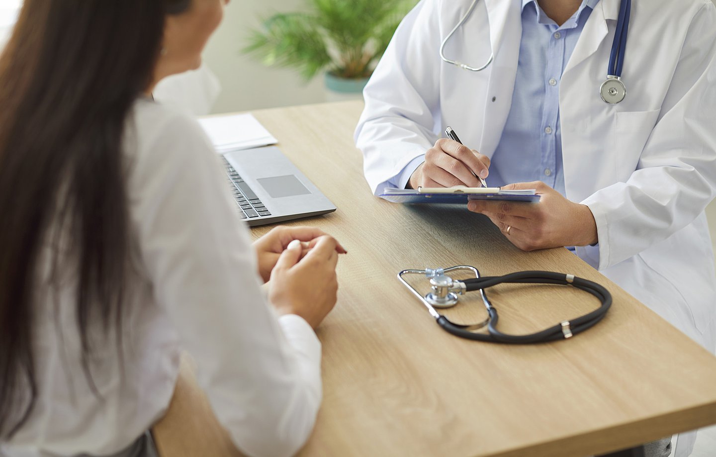 Ein Arzt sitzt im Gespräch mit einer Patientin am Schreibtisch des Behandlungszimmers. Auf dem Tisch liegt ein Stethoskop, in der Hand hält er ein Klemmbrett, in das er mit einem Stift Eintragungen vornimmt.