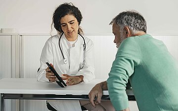 Eine &Auml;rztin ber&auml;t einen &auml;lteren Patienten, der ihr an einem Tisch gegen&uuml;bersitzt.