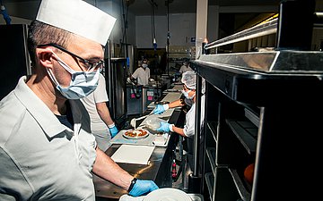 Foto von Küchenmitarbeitenden, die mit Maske Tabletts in einer Klinikkantine befüllen