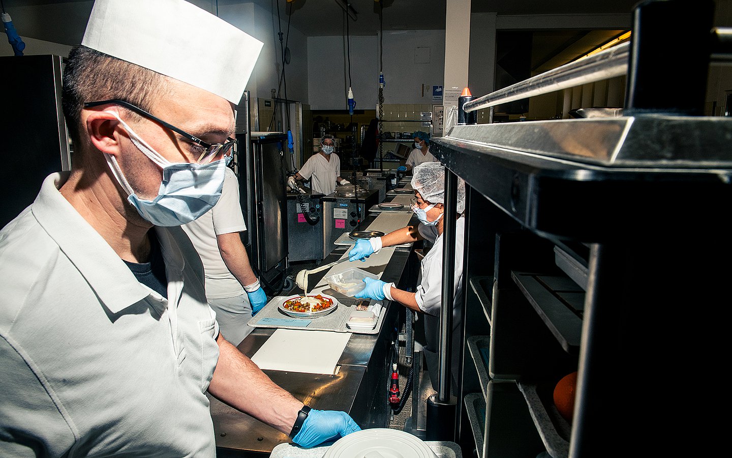 Foto von Küchenmitarbeitenden, die mit Maske Tabletts in einer Klinikkantine befüllen