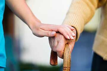 Eine Pflegerin ber&uuml;hrt eine pflegebed&uuml;rftige Person, die am Stock geht, sanft an der Hand