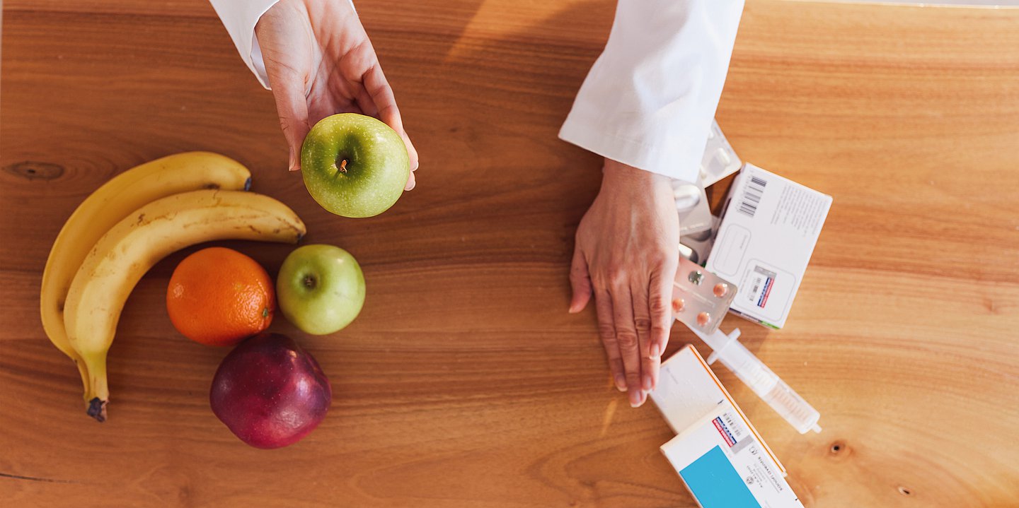 Foto von den Händen einer Medizinerin, die auf einem Tisch Tabletten zur Seite schieben und Obst anpreisen