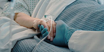 Eine &auml;ltere Dame liegt im Bett, die intraven&ouml;s &uuml;ber einen Katheter eine Infusion bekommt. Eine Pflegekraft h&auml;lt ihr die Hand.