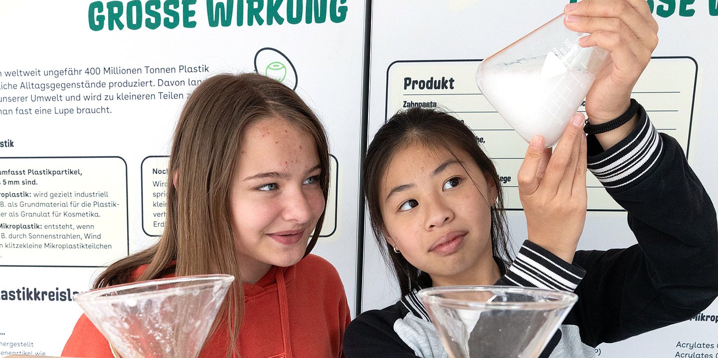 Zwei junge Mädchen führen ein Experiment zum Thema Mikroplastik durch. Sie stehen vor einer deutschen Informationstafel mit dem Titel 'GROSSE WIRKUNG', die über Plastikpartikel und deren Umweltauswirkungen informiert. Ein Mädchen in roter Jacke und eins in schwarz-weiß gestreiftem Pullover halten Glasschalen mit Wasser, während eine von ihnen eine weiße Substanz in das Wasser gibt. Die Tafel im Hintergrund erklärt verschiedene Arten von Plastikpartikeln und deren Entstehung durch Sonnenstrahlen, Wind und andere Umwelteinflüsse. Das Bild zeigt eine praktische Demonstration zur Veranschaulichung von Umweltbildung zum Thema Mikroplastik