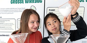 Zwei junge Mädchen führen ein Experiment zum Thema Mikroplastik durch. Sie stehen vor einer deutschen Informationstafel mit dem Titel 'GROSSE WIRKUNG', die über Plastikpartikel und deren Umweltauswirkungen informiert. Ein Mädchen in roter Jacke und eins in schwarz-weiß gestreiftem Pullover halten Glasschalen mit Wasser, während eine von ihnen eine weiße Substanz in das Wasser gibt. Die Tafel im Hintergrund erklärt verschiedene Arten von Plastikpartikeln und deren Entstehung durch Sonnenstrahlen, Wind und andere Umwelteinflüsse. Das Bild zeigt eine praktische Demonstration zur Veranschaulichung von Umweltbildung zum Thema Mikroplastik