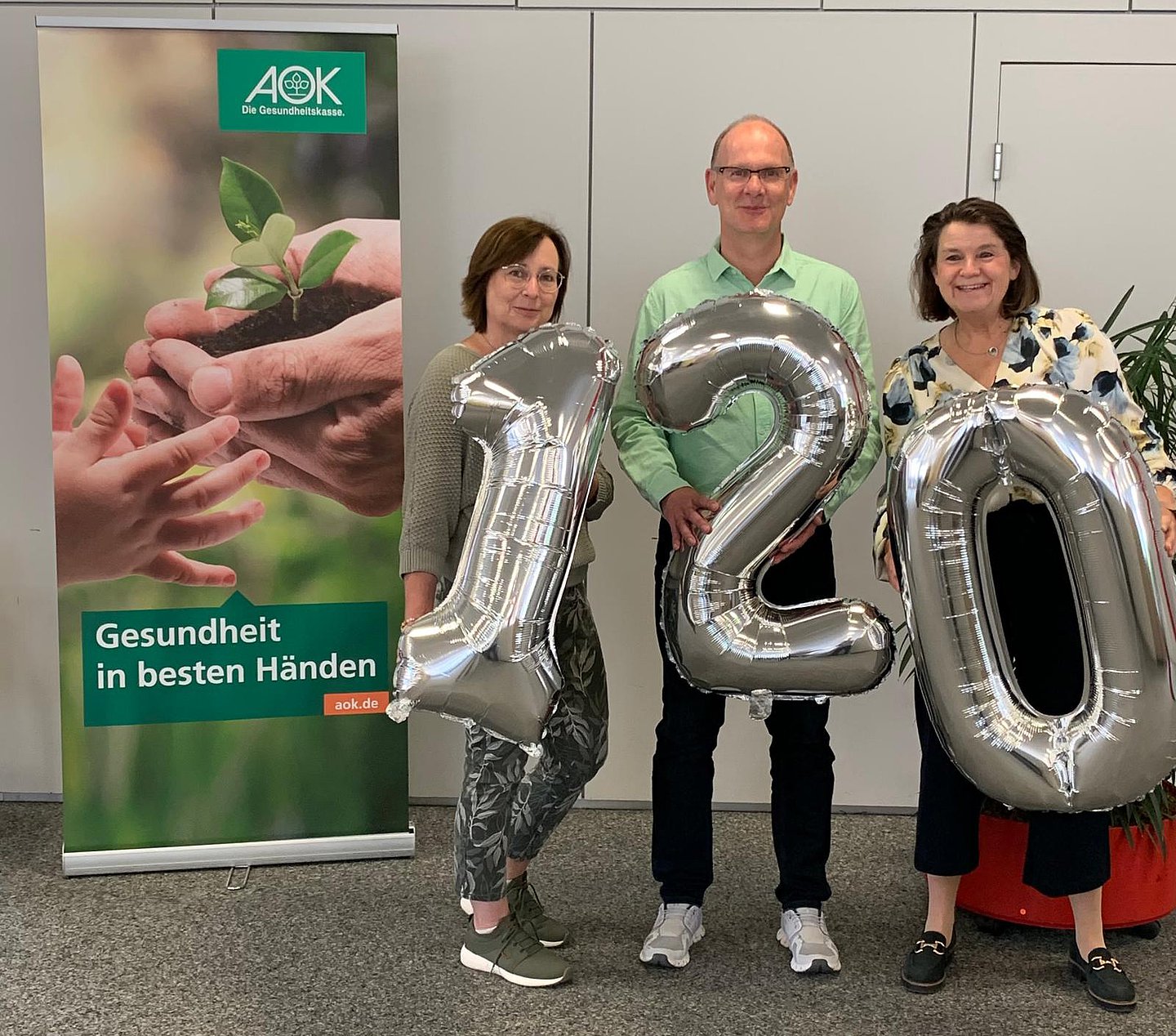 Das Foto zeigt einen Mann eingerahmt von zwei Frauen. Das Trio hält eine 120 aus silbernen Luftballons vor ihre Bäuche.