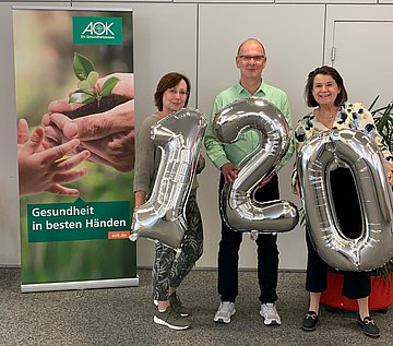 Gruppenbild 120 Jahre AOK Hessen Das Foto zeigt einen Mann eingerahmt von zwei Frauen. Das Trio hält eine 120 aus silbernen Luftballons vor ihre Bäuche.