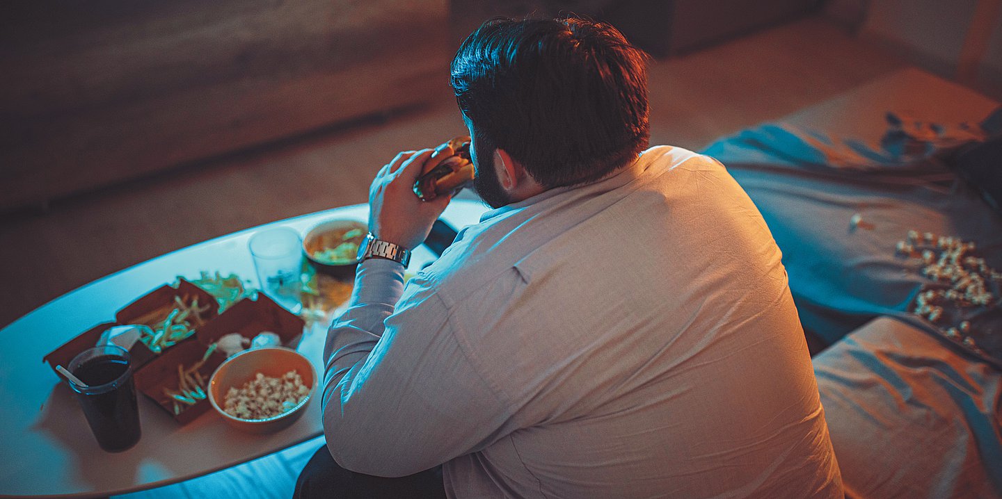 Kopfbild: Ein dicker Mann isst einen Burger auf der Couch, vor ihm auf dem Tisch verschiedene ungesunde Lebensmittel, wie Pommes, Popcorn, Chips und Cola..