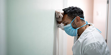Foto: Eine medizinische Kraft in wei&szlig;er Arbeitskleidung lehnt sich an die Wand und schaut gegen diese. 