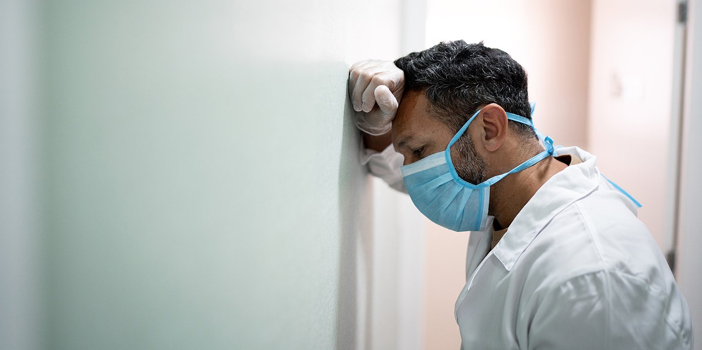 Foto: Eine medizinische Kraft in weißer Arbeitskleidung lehnt sich an die Wand und schaut gegen diese. 