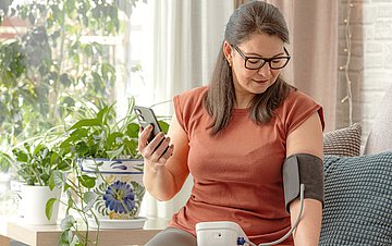 Online-Coach Bluthochdruck Foto einer Frau mittleren Alters, die im heimischen Wohnzimmer ihren Blutdruck misst.