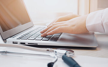 Foto: Zwei H&auml;nde schreiben auf der Tastatur eines Laptops, daneben liegt ein Stethoskop.