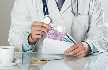 Ein Arzt, wei&szlig;er Kittel und ein Stethoskop um den Hals, sitzt an einem Tisch. In der linken Hand h&auml;lt er einen Briefumschlag, mit der rechten holt er einen 500-Euro-Schein hervor, weitere Geldscheine sind im Briefumschlag und auf dem Tisch zu sehen.