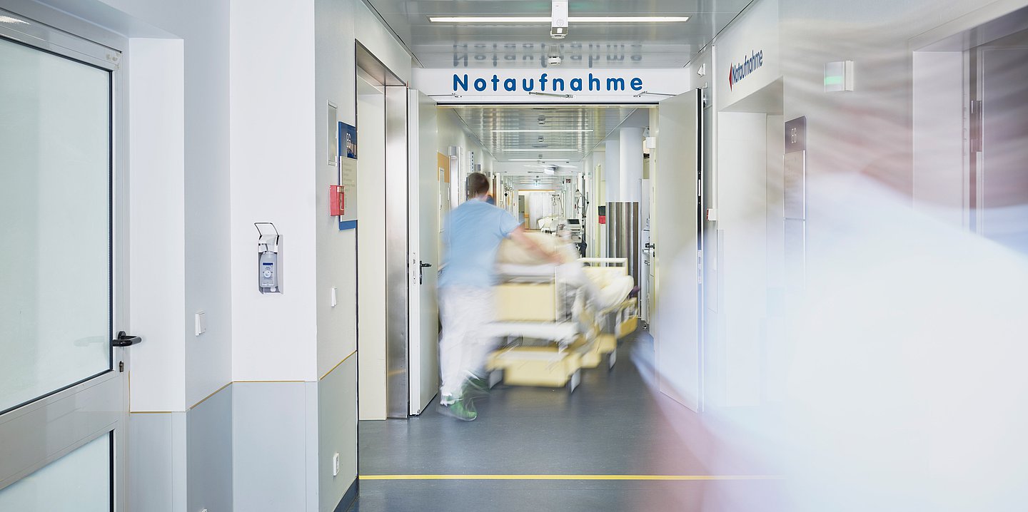 Ein Pfleger schiebt ein Bett den Flur der Notaufnahme eines Krankenhauses entlang.