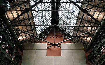 Foto: Industrieartig gestalteter Innenraum mit gro&szlig;er Glasdachkonstruktion, freiliegenden Stahltr&auml;gern und L&uuml;ftungsrohren sowie einer zentralen Wand unterhalb des Dachgeb&auml;lks.