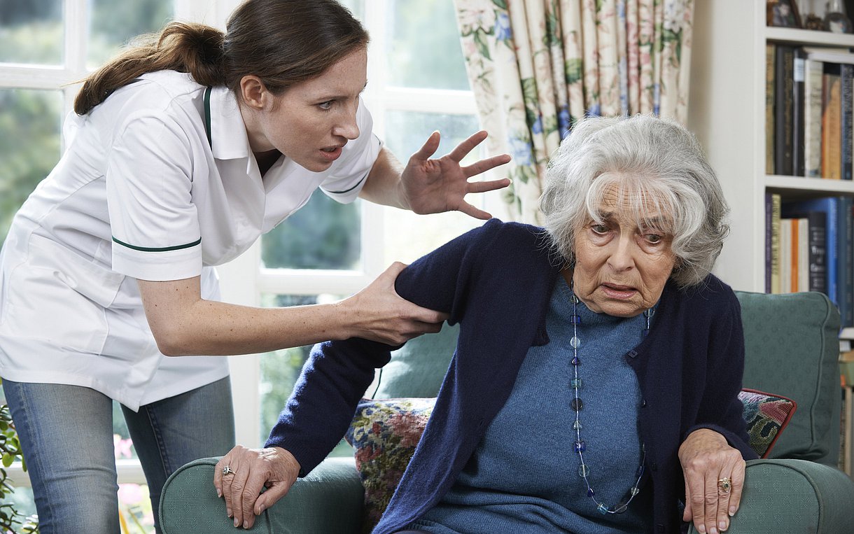 Foto: Eine Pflegerin greift eine ältere Dame am Arm und droht ihr mit erhobener Hand.