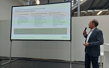Das Bild zeigt einen graumelierten Mann mit hoher Stirn und Brille. Er steht rechts unterhalb neben einem gro&szlig;en Bildschirm, auf dem eine Vortragspr&auml;snetation zu sehen ist. Der Mann tr&auml;gt einen graublauen Anzug und sein wei&szlig;es Hemd offen. Er blickt in Richtung Bildschirm und h&auml;lt in seiner rechten Hand ein schnurloses Mikrofon. 