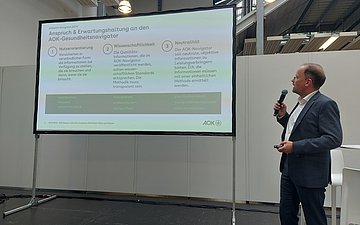 Das Bild zeigt einen graumelierten Mann mit hoher Stirn und Brille. Er steht rechts unterhalb neben einem gro&szlig;en Bildschirm, auf dem eine Vortragspr&auml;snetation zu sehen ist. Der Mann tr&auml;gt einen graublauen Anzug und sein wei&szlig;es Hemd offen. Er blickt in Richtung Bildschirm und h&auml;lt in seiner rechten Hand ein schnurloses Mikrofon. 