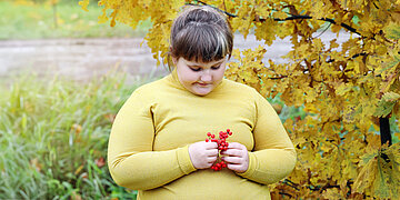 Ein adip&auml;oses M&auml;dchen in gelbem Pulli sitzt vor einem Busch in herbstlichen Farben und h&auml;lt rote Beeren in der Hand.