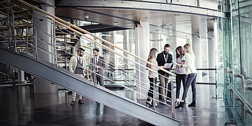  Das Bild zeigt den Treppenaufgang eines gl&auml;sernen B&uuml;rokomplexes. Rechts steht eine Gruppe aus einem Mann und drei Frauen zusammen und studiert Papiere. Auf der rechten Seite sind zwei M&auml;nner in ein Gespr&auml;ch vertieft.