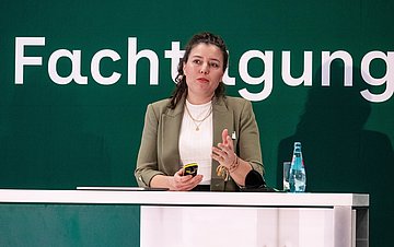 Auf dem Podium steht Uschi Jonas und hält einen Vortrag. Hinter ihr steht eine Wand mit der Aufschrift "Selbsthilfe-Fachtagung".