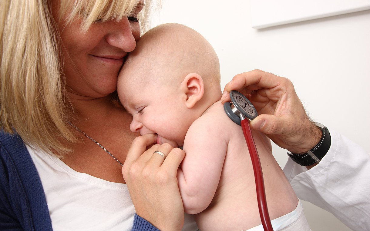Das Bild zeigt, wie ein Arzt ein Baby mit einem Stethoskop untersucht. Das Baby ist auf dem Arm einer Frau.