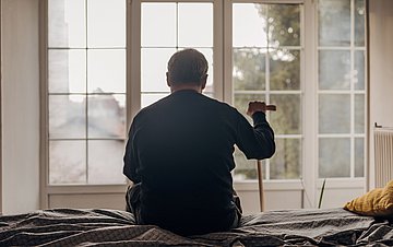 Altersdepression Foto eines alten Mannes mit Stock von hinten. Er sitzt auf einem Bett und blickt aus dem Fenster.