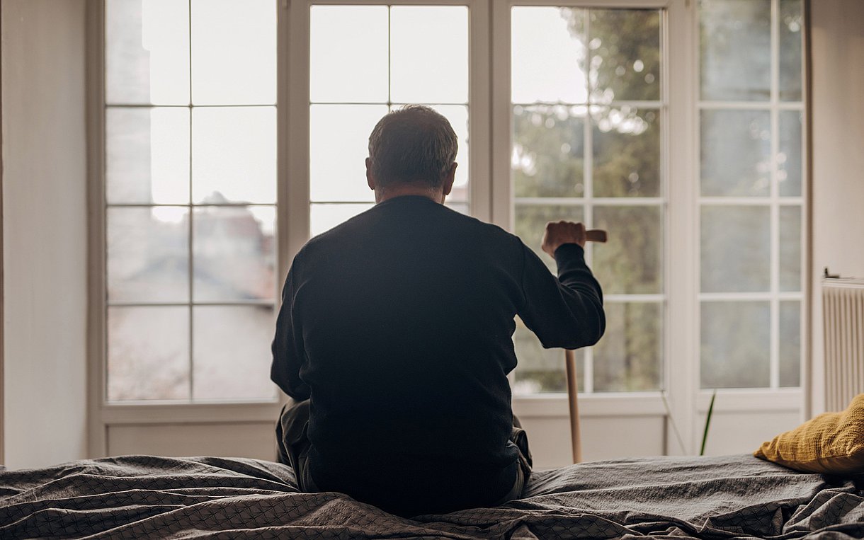 Foto eines alten Mannes mit Stock von hinten. Er sitzt auf einem Bett und blickt aus dem Fenster.