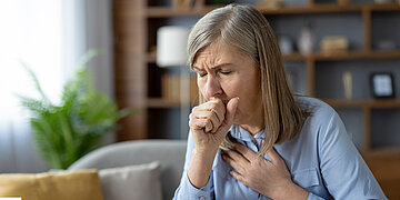 Eine &auml;ltere Frau h&auml;lt eine Hand auf die Brust und hustet in die andere Hand.