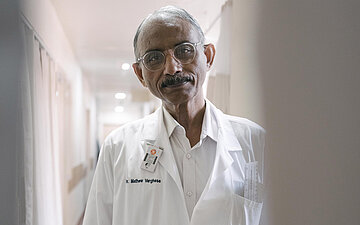 Dr. Mathew Varghese steht im Arztkittel im Flur und schaut in die Kamera. Er ist behandelnder Arzt und Spezialist f&uuml;r Klumpf&uuml;&szlig;e.