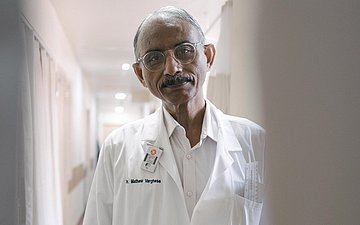 Dr. Mathew Varghese steht im Arztkittel im Flur und schaut in die Kamera. Er ist behandelnder Arzt und Spezialist für Klumpfüße.
