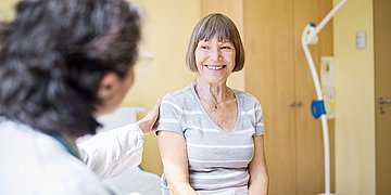 Ältere Frau lächelt im Patientenzimmer, eine Pflegekraft hält ihre Schulter.