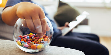 &Uuml;bergewichtiges Kind, das mit Tablet auf dem Sofa sitzt und mit der Hand in ein Glas Smarties greift.