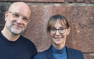 Portr&auml;t-Ansicht von Cornelia Koch und Boris Zirwes, die vor einer Ziegelwand stehen.