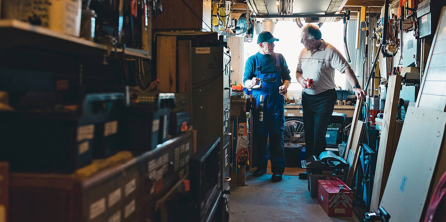 Foto: Zwei ältere Männer stehen in einer Werkstatt zusammen und reden miteinander.