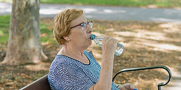 Eine Frau sitzt auf einer Bank im Park und trinkt Wasser aus einer kleinen Plastikflasche