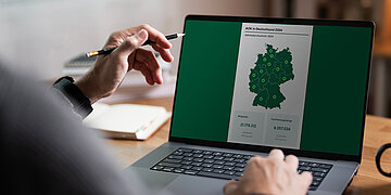 Ein Laptop steht auf einem Schreibtisch. Vor ihm eine Person, die mit einem Bleistift in der linken Hand. Sie schaut sich eine Grafik von Deutschland an, die die Versichertenzahl der (Mitglieder) und die Anzahl von Familienangehörigen in der GKV 2024 anzeigt.