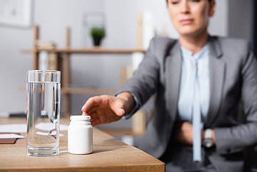 Magensäureblocker Westfalen-Lippe Eine Frau sitzt am Tisch, sich den Bauch haltend, und greift zu einer Packung Medikamenten.