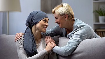 Förderung ambulante Hospizdienste in Schleswig-Holstein Eine junge Frau mit einem Tuch um den Kopf sitzt auf der Couch und wird von einer älteren Frau umarmt.