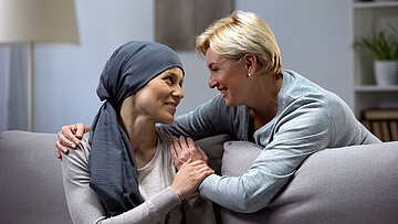 Eine junge Frau mit einem Tuch um den Kopf sitzt auf der Couch und wird von einer &auml;lteren Frau umarmt.