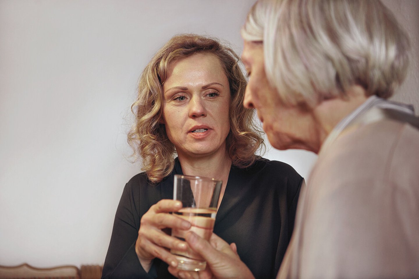 Eine Frau, vermutlich die Tochter, reicht einer Seniorin ein Glas Wasser.