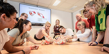 Um einem gro&szlig;en Tisch stehen viele Menschen und schauen auf ein Skelett in der Tischmitte. An diesem erkl&auml;ren zwei Frauen die Muskelbewegung des R&uuml;ckens. Im Hintergrund h&auml;ngt ein Monitor an der Wand, auf dem unterst&uuml;tzend die Muskeln abgebilldet sind.