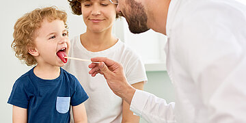 Ein kleines Kind mit lockigen Haaren sitzt in einem Untersuchungsraum und streckt die Zunge heraus, w&auml;hrend ein Arzt mit einem Holzspatel den Hals untersucht. Ein Elternteil sitzt daneben und schaut zu.