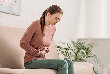 Magensäureblocker Schleswig-Holstein Eine junge Frau sitzt leicht vornübergebeugt auf einem Sofa im Wohnzimmer und hält sich mit schmerzverzerrtem Gesicht beide Hände vor dem Oberbauch.