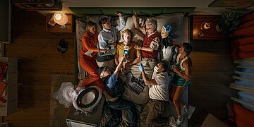 Das Bild zeigt eine Gruppe verschiedener Personen auf einem Doppelbett von oben, die nicht alle schlafen, sondern sich mit anderen Dingen besch&auml;ftigen