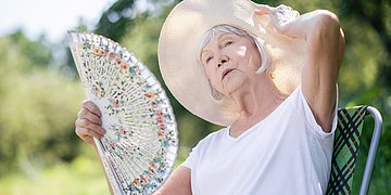 Gefährliche Hitze Eine ältere Dame sitzt in der Natur im Schatten. Sie trägt einen Hut und fächert sich mit einem mit Blumen bemalten Fächer Luft zu.